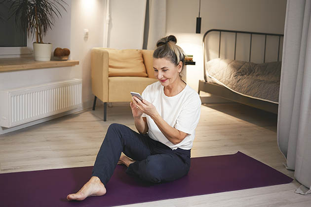 Health, fitness, vitality, age and modern technology. Cheerful athletic mature female in sports clothes sitting on yoga mat, smiling, checking calorie loss using app on cell phone after training