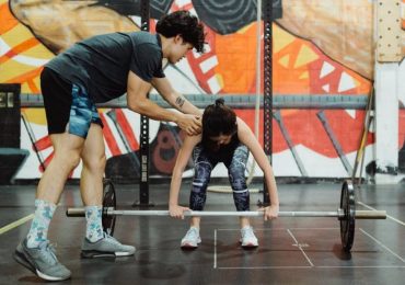 A gym owner helps their student through a beginner weight lifting exercise.