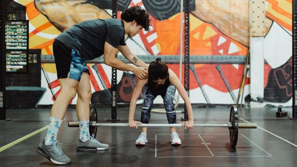 A gym owner helps their student through a beginner weight lifting exercise.
